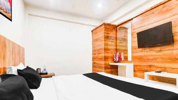 A hotel room with a bed, a flat-screen TV mounted on the wall, a wooden wardrobe, a small bedside table with a vase of flowers, and a water bottle on the nightstand. - Collection O Indore Gandhi Hall (Indore)