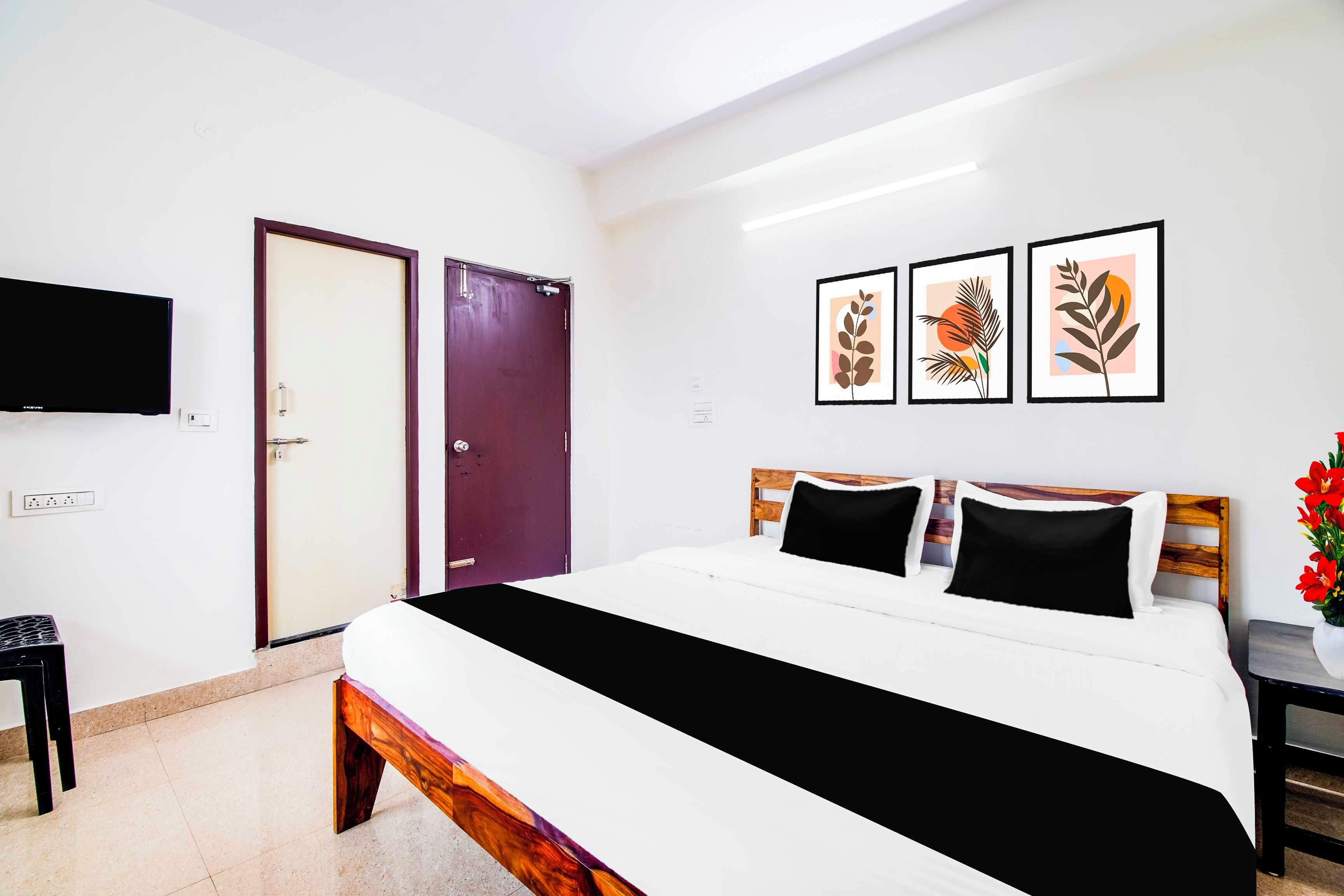 A hotel room with a bed, a TV, a door, and three framed artworks on the wall.