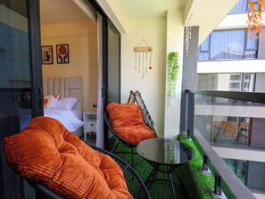 A balcony with a bed, orange cushions, a glass table, and a hanging plant. - Modern 1-Bedroom near Sarit Centre Mall, Museum, City attractions & Top Dining (Nairobi)