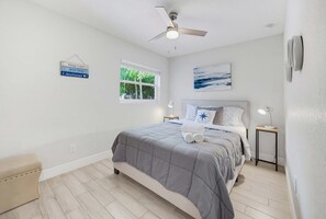 Ein Schlafzimmer mit Bett, Deckenventilator, Fenster, Nachttisch mit Lampe und einem Wandbild.