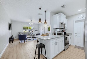A modern kitchen with white cabinetry, a stainless steel microwave, and a built-in oven. There is a bar area with stools and a seating area with a grey sofa and a blue chair. - Fenced Backyard 2BR Pet Friendly and Sleeps 6 (Clearwater)
