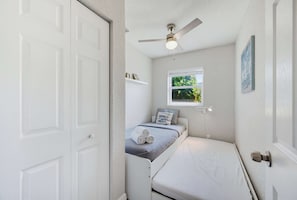 Habitació compacte amb un llit individual, un ventilador de sostre, una finestra amb vista a vegetació i una petita estant amb objectes.