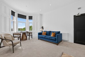 Ein Zimmer mit einem blauen Sofa, einem Esstisch mit Stühlen und großen Fenstern.