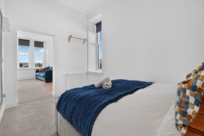 A bedroom with a bed, a window, a radiator, and a view of a blue sofa in the adjacent room. - Seaview Largs - 1 Bedroom Ground Floor Apartment (Largs)