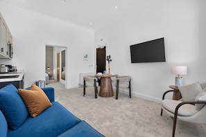 A modern living room with a blue sofa, a dining table with chairs, a flat-screen TV mounted on the wall, and a small side table with a lamp. - Seaview Largs - 1 Bedroom Ground Floor Apartment (Largs)