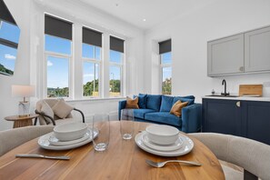 A modern living room with a dining table set for two, a blue sofa, and a kitchen area with a sink and cabinets. - Seaview Largs - 1 Bedroom Ground Floor Apartment (Largs)
