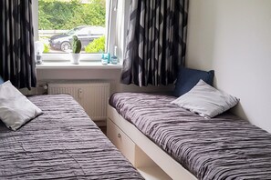 Ein Zimmer mit zwei Betten, einem Heizkörper, einem Fenster mit Vorhängen und Blick auf einen parkenden Wagen vor dem Haus.