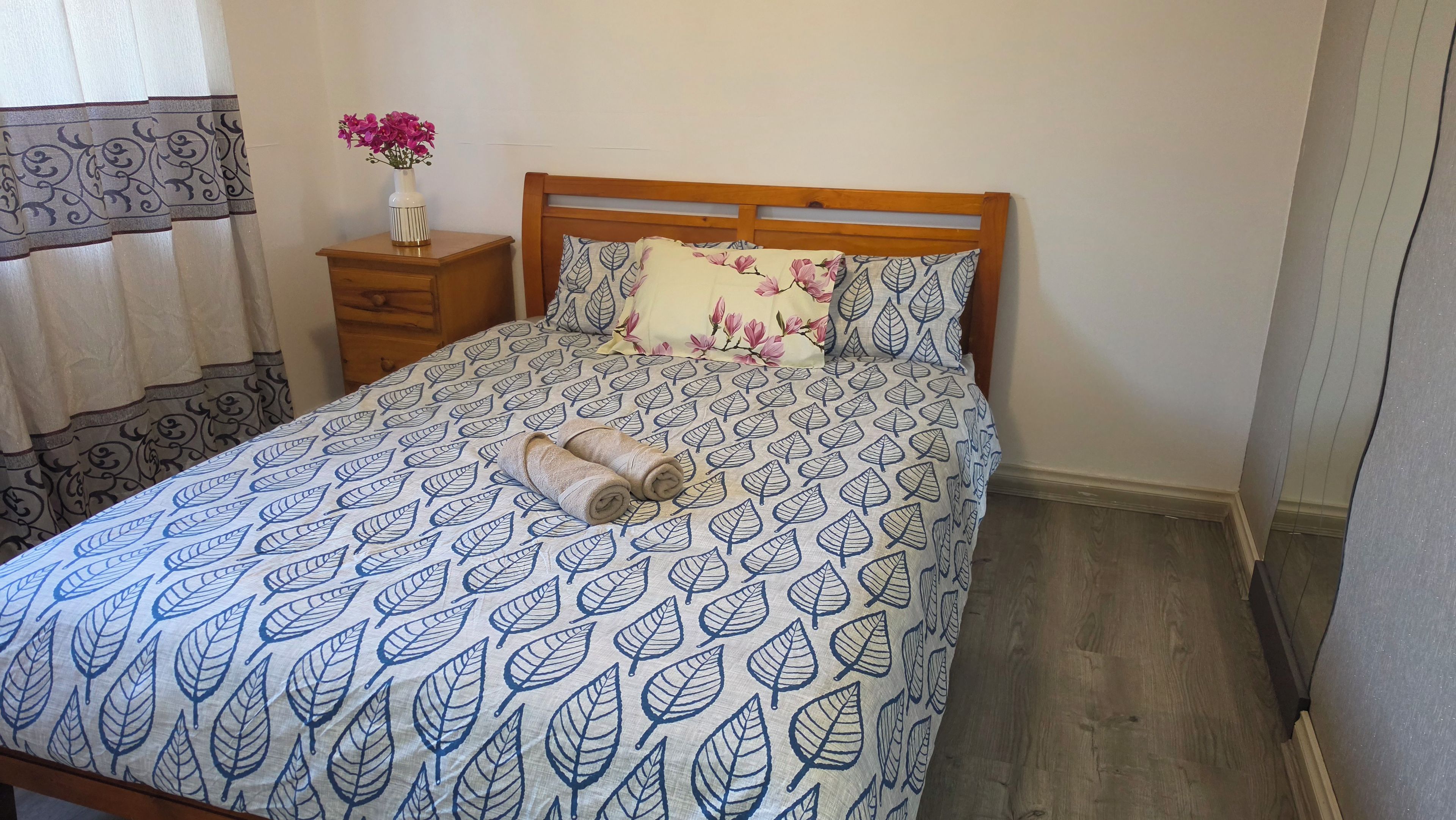A bed with a leaf pattern bedspread, a wooden headboard, a nightstand with a vase of flowers, and a pillow with a floral design.