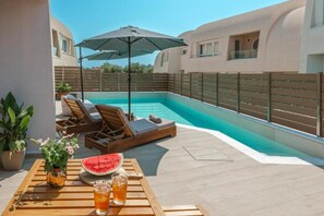 Ein Poolbereich mit Liegestühlen, einem Tisch mit Getränken und einer Wassermelone, sowie einem großen Sonnenschirm.