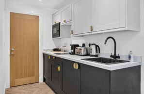 A modern kitchen with a wooden door, white cabinets, a black sink, and various appliances. - Seaview Largs - 1 Bedroom Apartment (Largs)