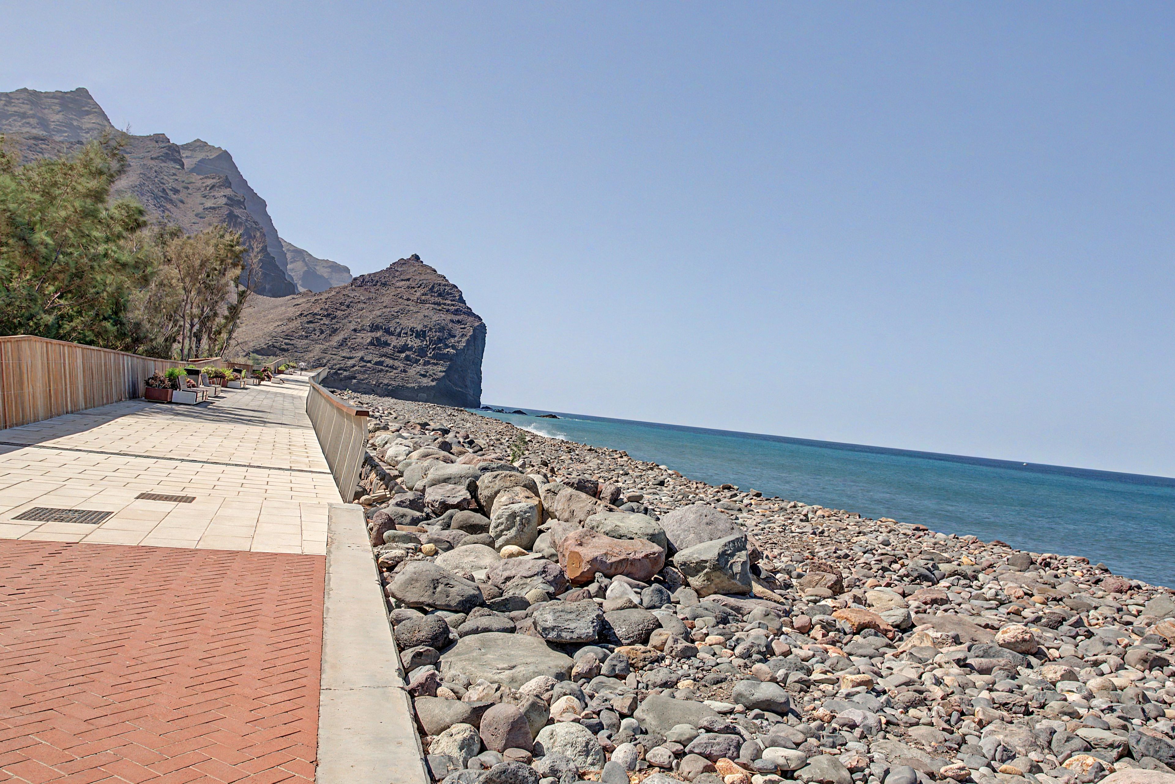 A paved walkway beside a rocky shoreline with a mountain in the background.