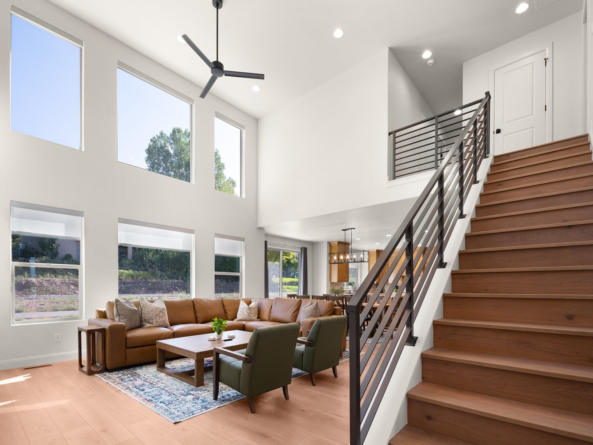Amplia sala de estar con techo alto, ventanas grandes, una escalera y una cocina moderna al fondo.