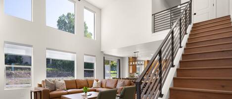 Amplia sala de estar con techo alto, ventanas grandes, una escalera y una cocina moderna al fondo.