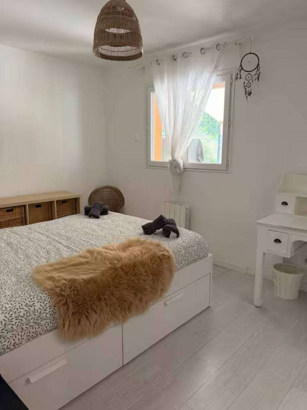 Een slaapkamer met een bed, een wit bureau, een raam met gordijnen en een dromenvanger aan de muur.