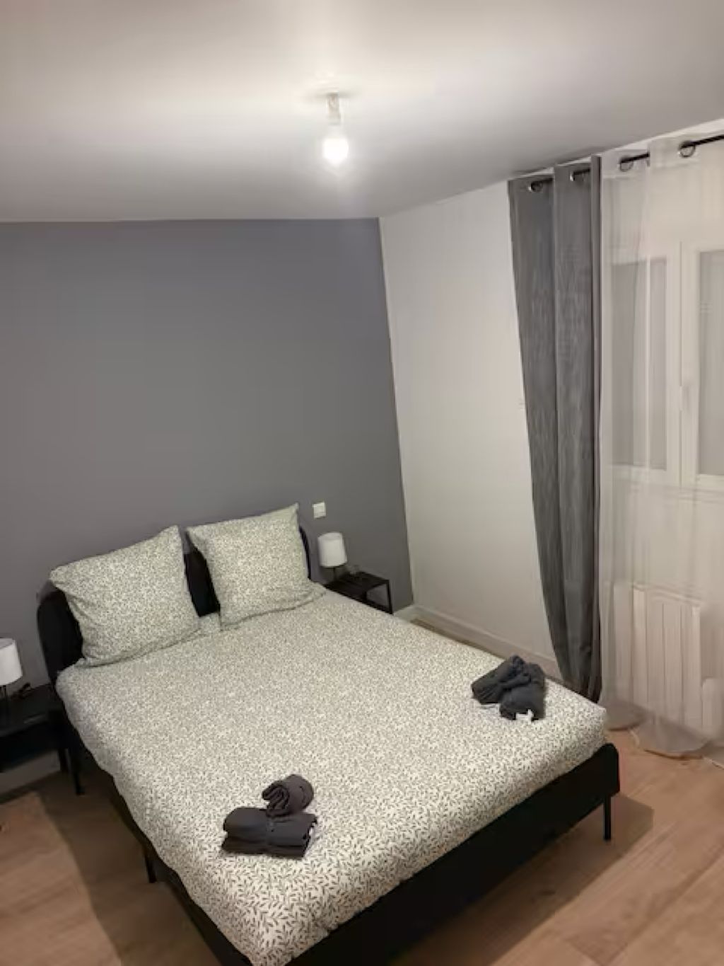 Een eenpersoonsbed met een bedsprei met dessin, twee kussens, een nachtkastje met een lamp en een raam met gordijnen.