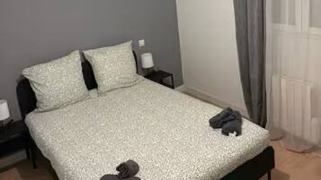 Een eenpersoonsbed met een bedsprei met dessin, twee kussens, een nachtkastje met een lamp en een raam met gordijnen.