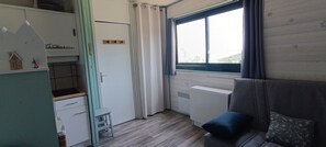 A compact room with a kitchenette, a small stool, a window with curtains, and a sofa with a cushion. - Cozy studio apartment 50 meters from the base of the slopes (Arette la Pierre saint)