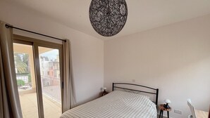 Een slaapkamer met een bed, nachtkastje, stoel, tafel en balkon met gordijnen.