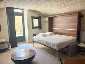 A bedroom with a bed, a round table, and a view of the outside. - Stone studio apartment, downtown Uzès, large inner courtyard: La Capitelle (Uzès)