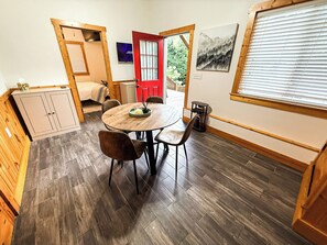 A room with a round wooden table, two chairs, a bed, a red door, and a window with blinds. - Tree Top Retreat - Where Mountain Luxury Meets Adventure (Fleetwood)