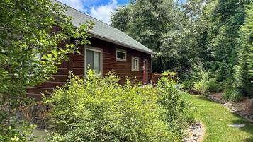 A wooden cabin with a stone foundation, surrounded by dense greenery and trees.