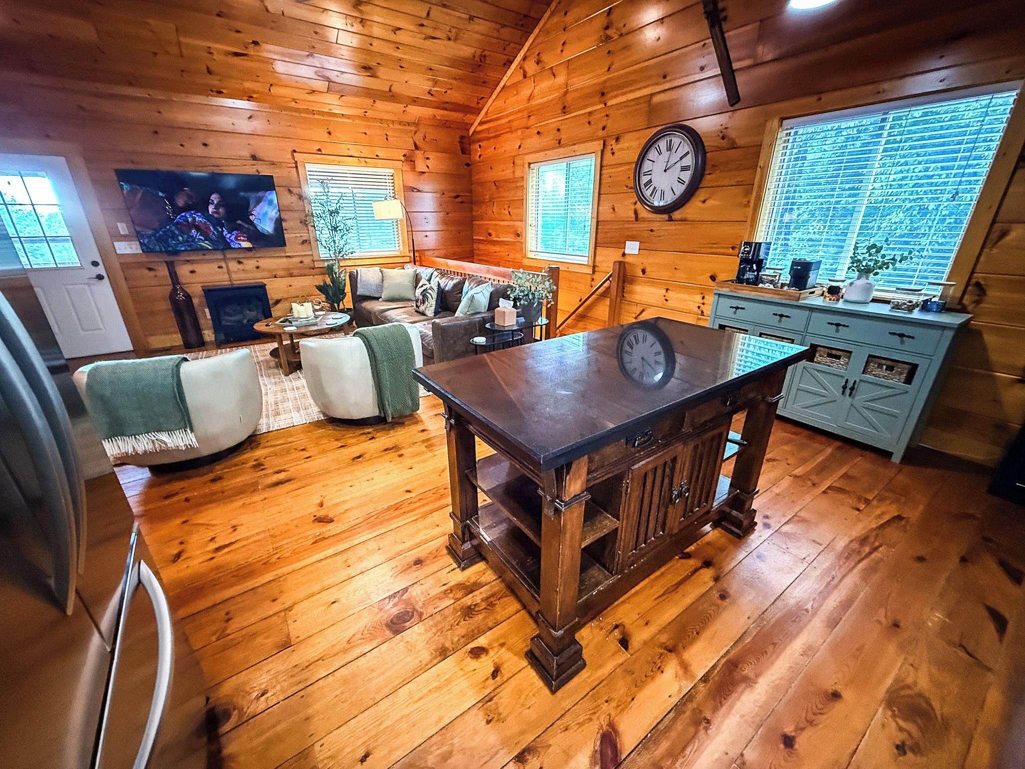 A cozy cabin interior with a wooden floor, a dining table, a sofa, a television, and a kitchen area with a clock.