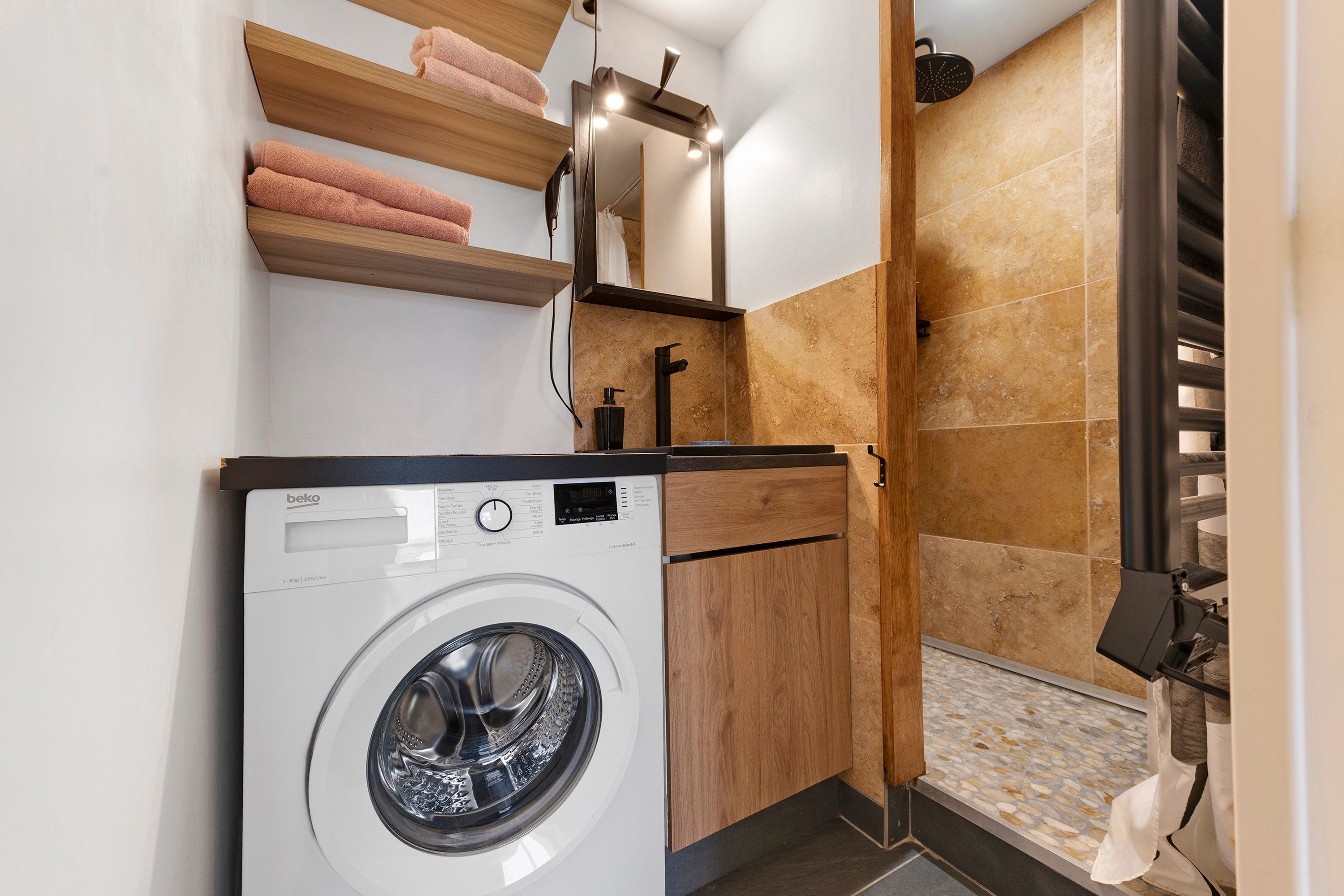 Un espacio de lavandería compacto con lavadora de carga frontal, estantes de madera con toallas, un espejo y una zona de ducha con pared azulejada.