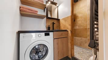 Un espacio de lavandería compacto con lavadora de carga frontal, estantes de madera con toallas, un espejo y una zona de ducha con pared azulejada.