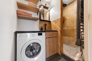 A compact laundry area with a front-loading washing machine, wooden shelves with towels, a mirror, and a shower area with a tiled wall.