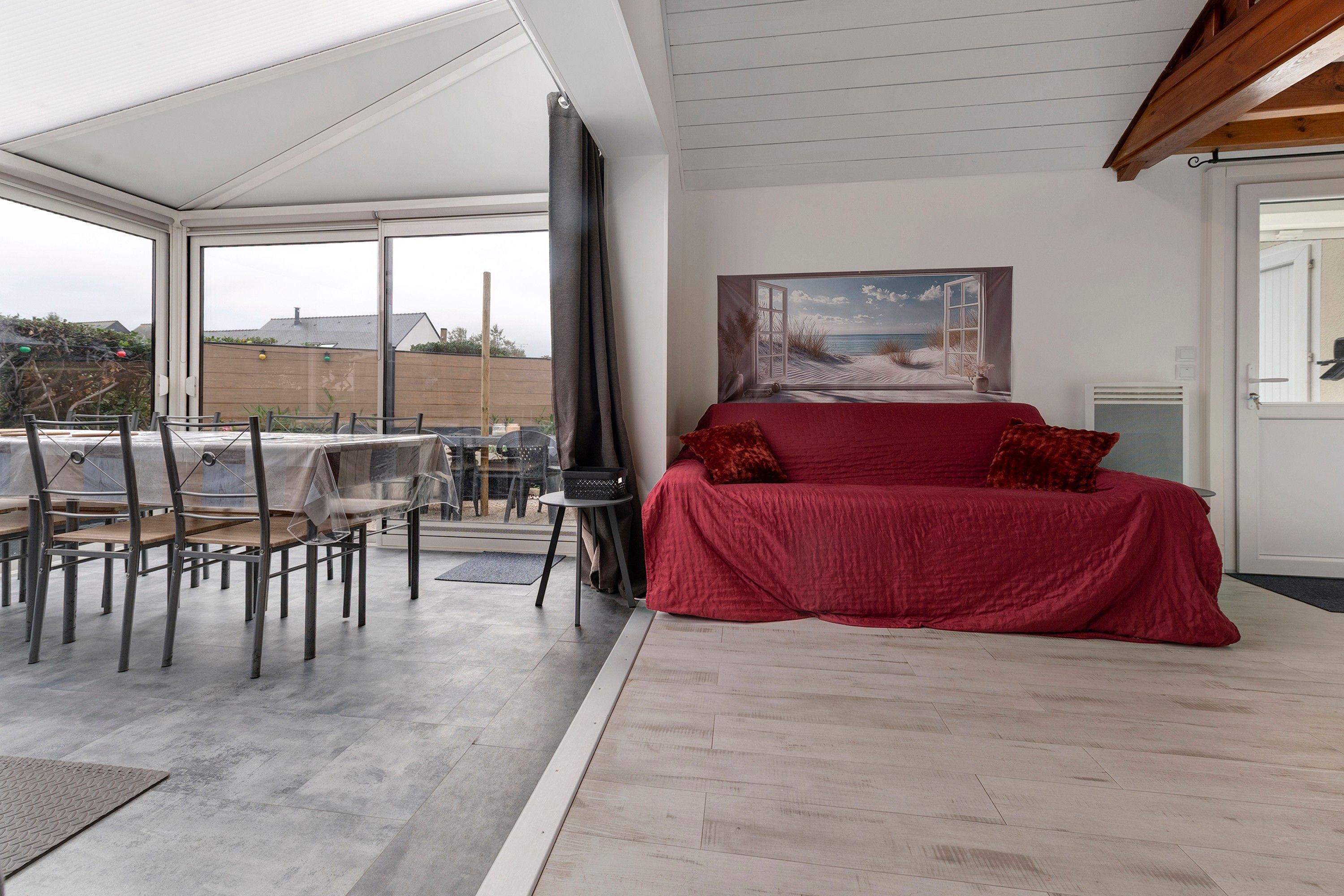 A modern living room with a red sofa, a painting of a beach scene, and a view of the outdoors.