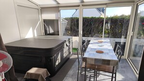 A sunlit room with a hot tub, a table with chairs, and a view through large windows. - Holiday Home 'Le Cottage Des Dunes - Erdeven' with Private Terrace, Garden and Wi-Fi (Erdeven)
