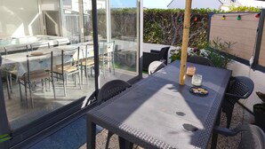 A glass-enclosed patio with a table and chairs, a view of the interior, and a wooden fence with plants. - Holiday Home 'Le Cottage Des Dunes - Erdeven' with Private Terrace, Garden and Wi-Fi (Erdeven)
