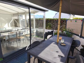 A glass-enclosed patio with a table and chairs, a view of the interior, and a wooden fence with plants. - Holiday Home 'Le Cottage Des Dunes - Erdeven' with Private Terrace, Garden and Wi-Fi (Erdeven)