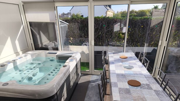 A hot tub and a covered dining table with chairs, set on a balcony with a view of a garden and buildings. - Holiday Home 'Le Cottage Des Dunes - Erdeven' with Private Terrace, Garden and Wi-Fi (Erdeven)