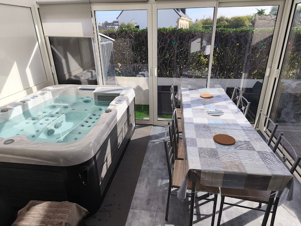 A hot tub and a covered dining table with chairs, set on a balcony with a view of a garden and buildings. - Holiday Home 'Le Cottage Des Dunes - Erdeven' with Private Terrace, Garden and Wi-Fi (Erdeven)