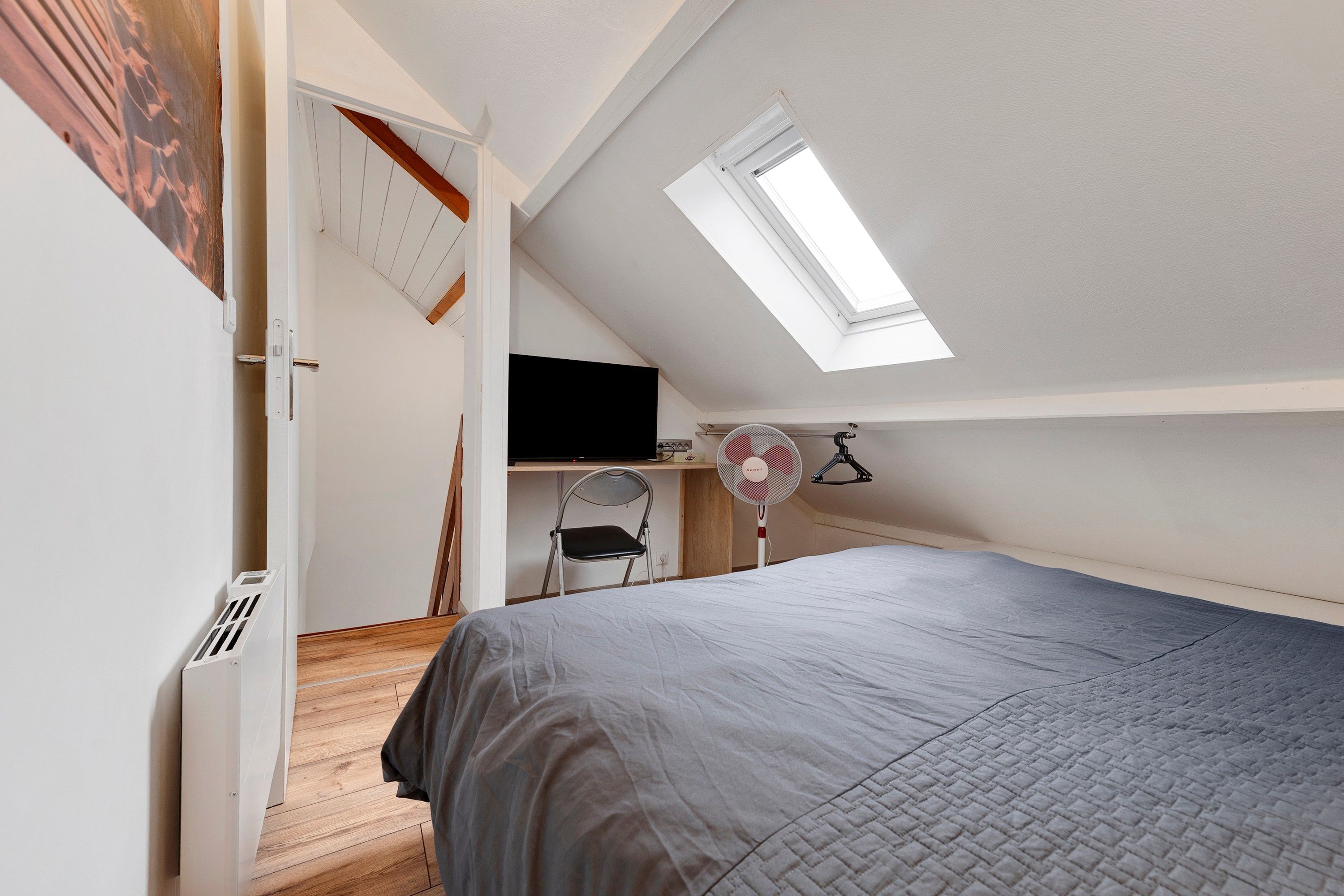 A bedroom with a sloped ceiling, a skylight, a bed, a chair, a desk, and a wall-mounted fan.