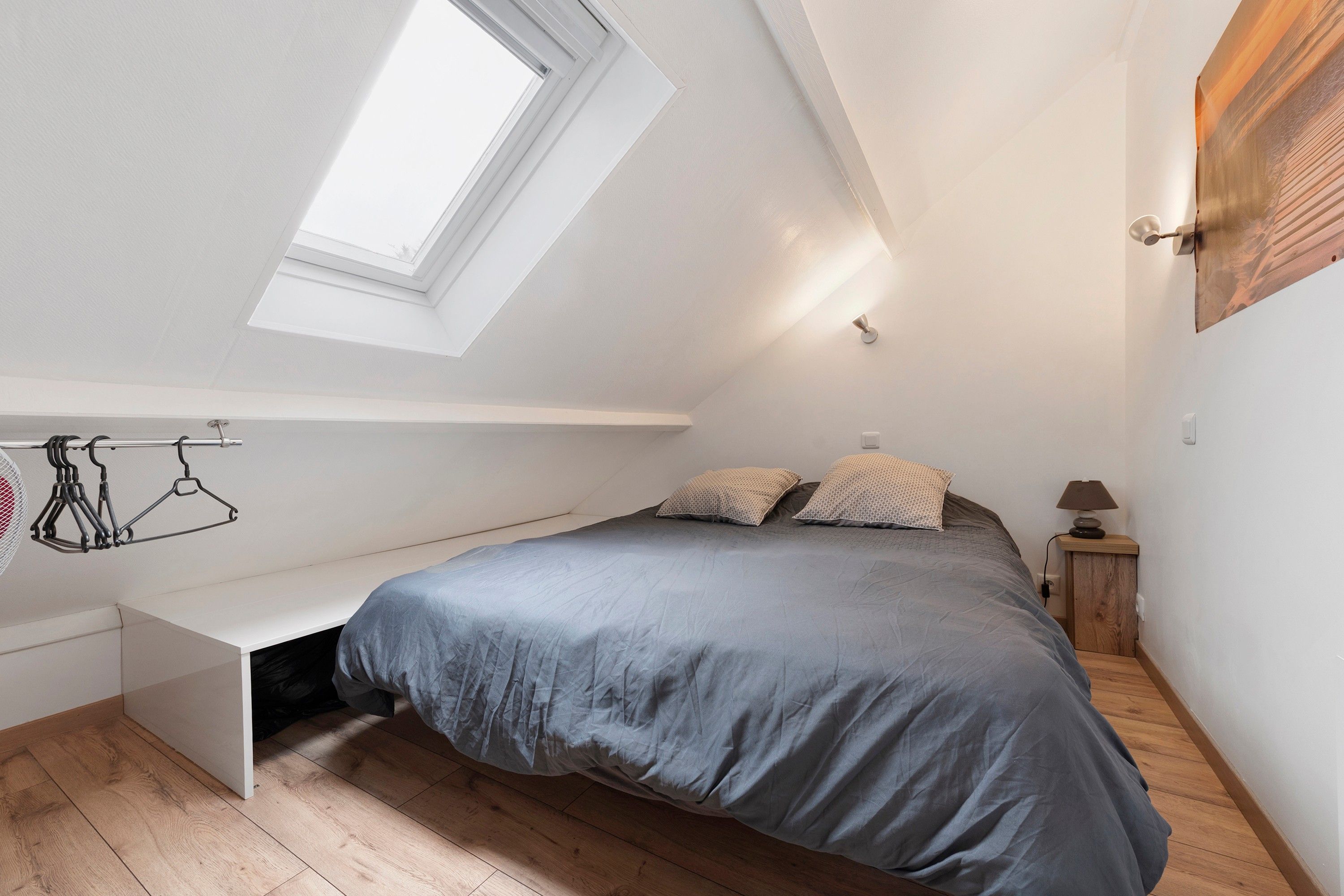 Une chambre avec un puits de lumière, un lit avec des oreillers, une table de chevet avec une lampe et un porte-manteau fixé au mur.