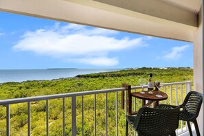 A balcony with a table and chairs overlooking a coastal landscape with greenery and the sea. - Key Lime Sunrise Top Floor Oceanfront 3404 (Tavernier)