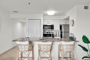 A modern kitchen with white cabinets, stainless steel appliances, a marble countertop, and three wicker bar stools. - Key Lime Sunrise Top Floor Oceanfront 3404 (Tavernier)