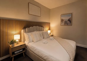 Ein ordentlich bezogenes Bett mit Kissen und Decke, ein Nachttisch mit Lampe und Buch sowie eine Wand mit vertikalen Holzlatten.