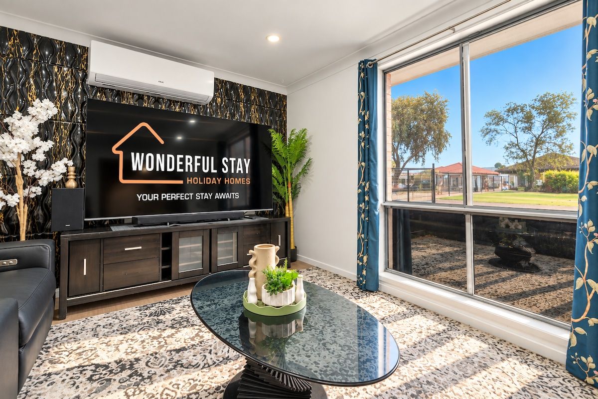 A modern living room with a large flat-screen TV, a glass coffee table, and a view of the outdoors.