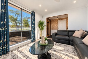 A modern living room with a glass coffee table, a black leather sofa, and a view of a fenced outdoor area.