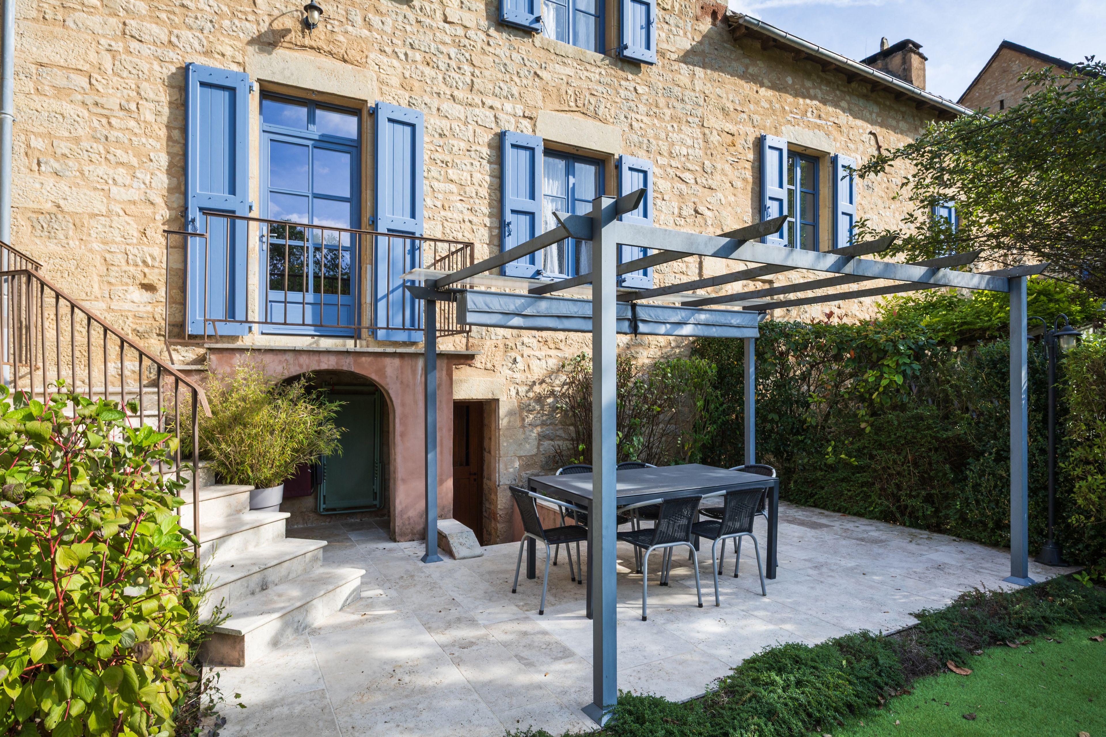 Un bâtiment en pierre avec des volets bleus, un patio couvert avec une table et des chaises, et un escalier menant à l’entrée.
