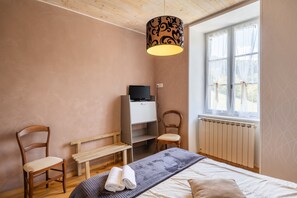 A bedroom with a bed, a desk, two chairs, a bench, a radiator, and a window with curtains. - Lou Flaujer Cottage: Mountain View, Private Terrace & Wi-Fi in Pont-les-Bains (Salles-la-Source)