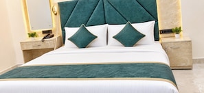 A bed with a tufted headboard, two pillows, a bedside table with a phone, and a nightstand with a plant. - The Rama Inn (Faizabad)