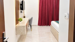 A hotel room with a bed, a desk with a chair, a small plant, and a red curtain. - The Rama Inn (Faizabad)