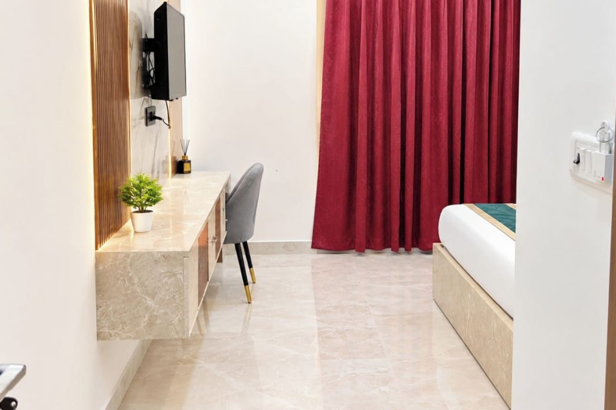 A hotel room with a bed, a desk with a chair, a small plant, and a red curtain.