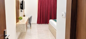 A hotel room with a bed, a desk with a chair, a small plant, and a red curtain. - The Rama Inn (Faizabad)