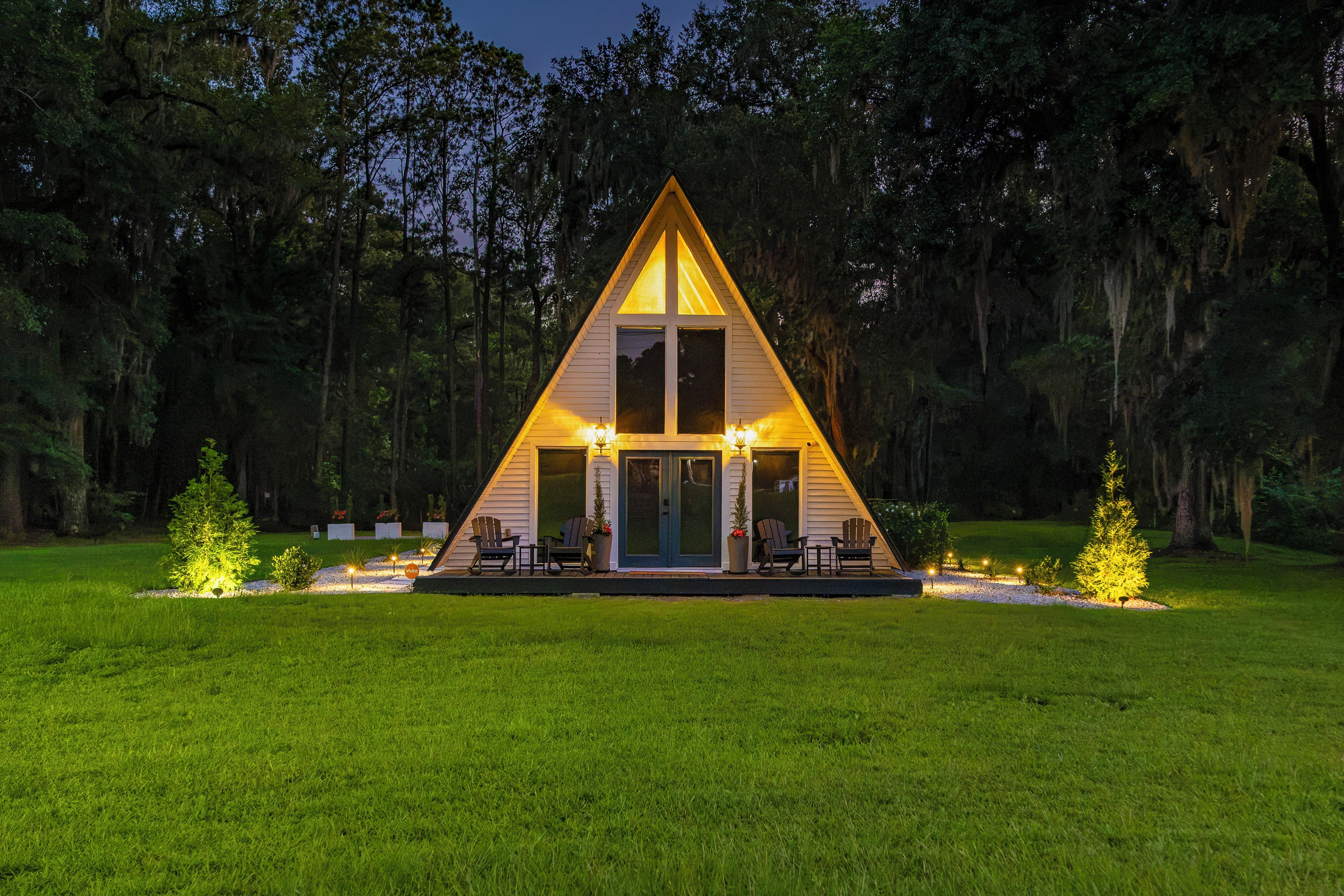 Une cabane de forme triangulaire, dotée d’un porche et entourée d’une zone herbeuse et d’arbres.