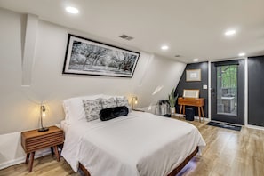 A bedroom with a bed, bedside tables, a wall-mounted picture, a door with a view, and a small wooden side table.